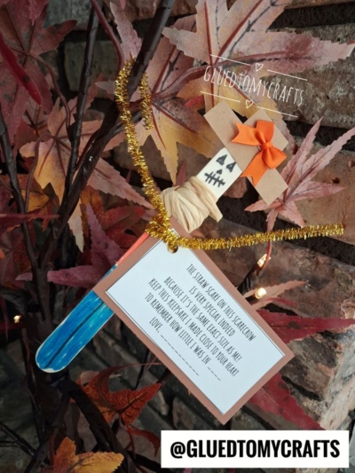 A popsicle stick craft shaped like a spooky knife with a jack-o-lantern face, orange bow, and message card hangs on fall leaves; this fun scarecrow height ornament adds festive charm to your autumn decor—labeled @GLUEDTOMYCRAFTS.