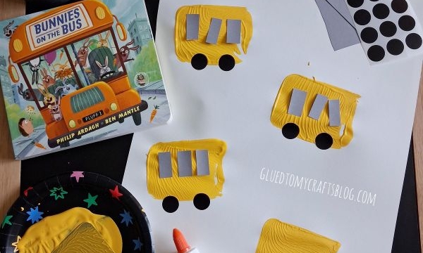 Children’s craft setup with yellow paint, glue, black dot stickers, paper buses, a “Turkey Goes to School” book, “Bunnies on the Bus” book, and craft supplies in metal cups on a dark surface.