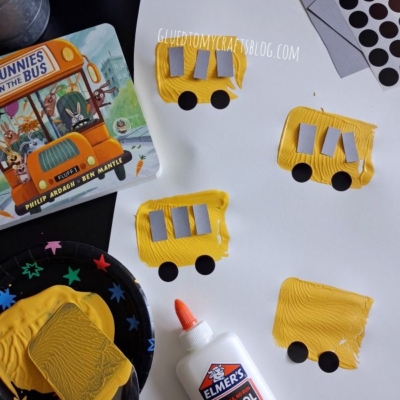 Children’s craft project: yellow painted rectangles on white paper with black wheels and gray windows, resembling school buses. Supplies include glue, paint, a sponge, stickers, and a “Bunnies on the Bus” book.