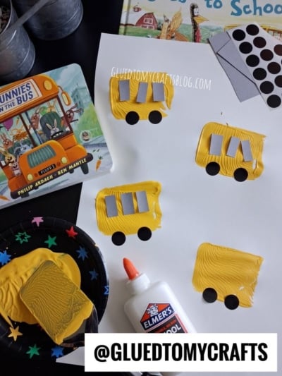 Children’s craft project: yellow painted rectangles on white paper with black wheels and gray windows, resembling school buses. Supplies include glue, paint, a sponge, stickers, and a “Bunnies on the Bus” book.