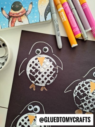 A penguin craft using white paint, googly eyes, and orange paper on black paper, with markers and a painted penguin card nearby. @GLUEDTOMYCRAFTS is written at the bottom.