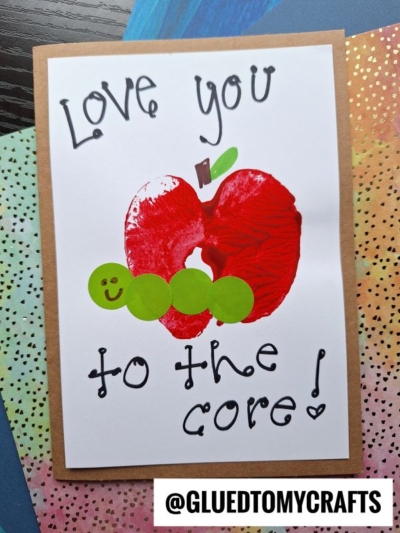 Handmade card with a charming red apple print in an apple stamped craft style, a green caterpillar made of paper circles, and the handwritten message Love you to the core! on white paper. @GLUEDTOMYCRAFTS is printed at the bottom.