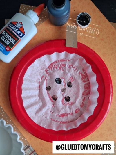 A red plate holds a coffee filter with holes and paint marks, perfect for FALL CRAFTS FOR KIDS. Nearby are a bottle of school glue, a blue paint bottle, and orange patterned paper. @GLUEDTOMYCRAFTS is written at the bottom.