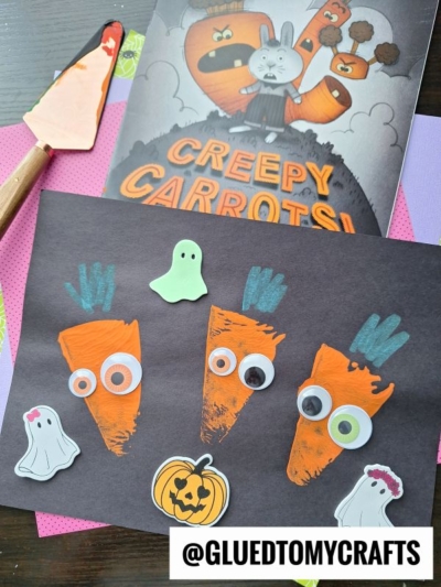 Children’s artwork inspired by Creepy Carrots! shows three painted orange carrots with googly eyes on black paper, surrounded by ghost and pumpkin stickers. The book, a carrot-shaped spatula, and a pie server stamped with a playful message are nearby.