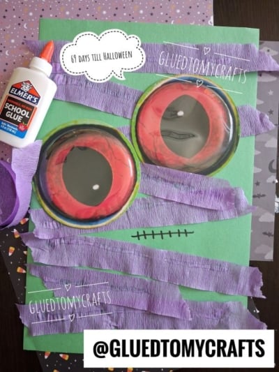 Colorful Halloween craft scene features large red monster eyes, purple crepe paper strips, green paper, Elmers glue, and patterned purple paper—perfect inspiration for Halloween Crafts for Kids. A speech bubble says “61 days till Halloween.” @GluedToMyCrafts is shown.