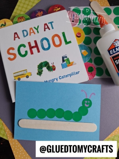 Children’s craft supplies and book. A blue card with a green caterpillar made of dot stickers and a popsicle stick is next to The Very Hungry Caterpillar school book, Elmer’s glue, and colorful coding label crafts. Green sticker dots are scattered nearby.