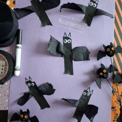 Six handmade bat crafts made from black paper and googly eyes are arranged on a purple sheet, along with a marker and Halloween-themed decorations. A white banner reads @GLUEDTOMYCRAFTS..