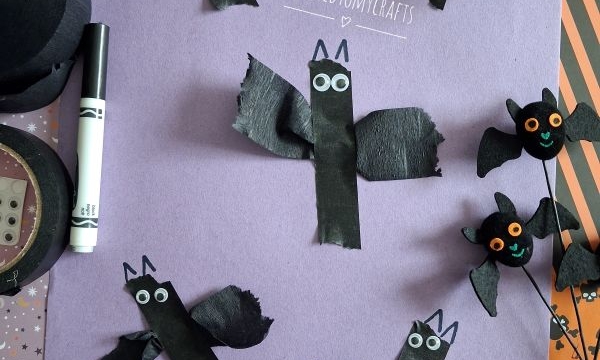 Six handmade bat crafts made from black paper and googly eyes are arranged on a purple sheet, along with a marker and Halloween-themed decorations. A white banner reads @GLUEDTOMYCRAFTS..