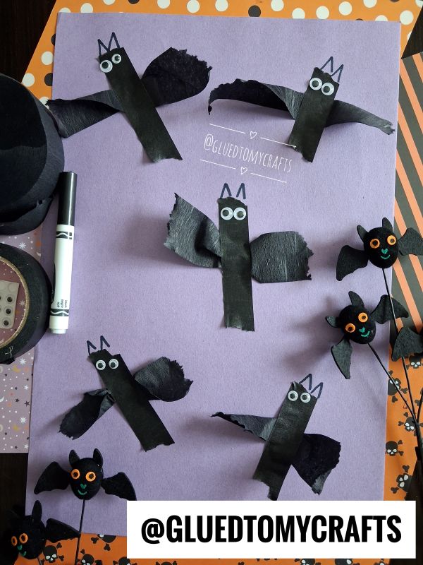Six handmade bat crafts made from black paper and googly eyes are arranged on a purple sheet, along with a marker and Halloween-themed decorations. A white banner reads @GLUEDTOMYCRAFTS..