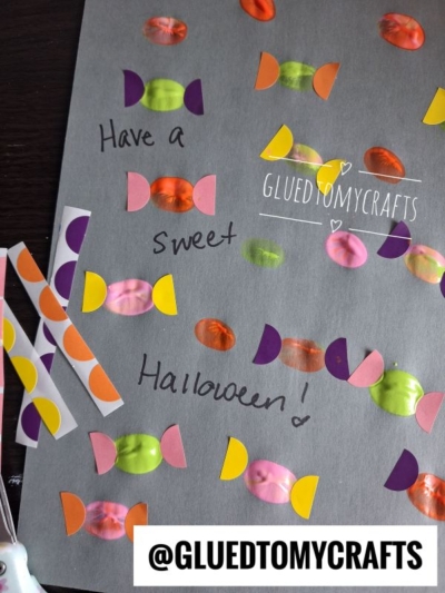 A gray card decorated with colorful candy stickers and text that says, Have a sweet Halloween! Perfect for Halloween Crafts for Kids, the @GLUEDTOMYCRAFTS handle is at the bottom on a white strip.