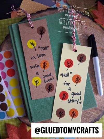 Two handmade bookmarks with leaf stickers and handwritten Fall-themed messages rest on a green book, making perfect fall crafts for kids. A marker and colorful dot stickers are nearby. One tag reads Fall in love with a good book! The other, Fall for a good story!.