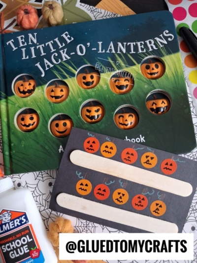 A Halloween craft scene perfect for halloween story time, featuring a Ten Little Jack-O-Lanterns book, a pumpkin sticker card using craft sticks, Elmer’s glue, and colorful paper—all set on a spiderweb-patterned background.