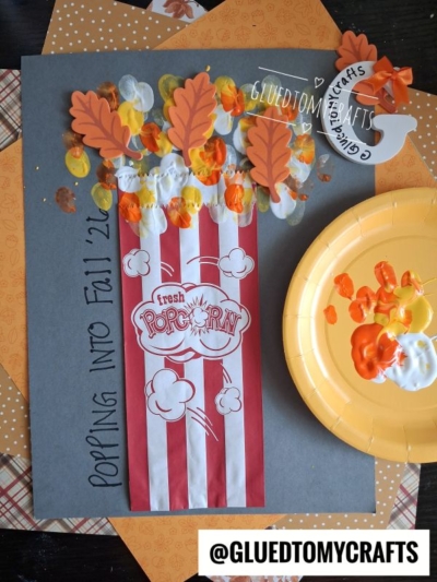 A fall-themed popcorn craft—perfect for FALL CRAFTS FOR KIDS—features red-and-white stripes, paper leaves, and painted yellow, orange, and white popcorn on gray paper. The words “Popping Into Fall 22” are written on the side. A paint-covered plate sits nearby.
