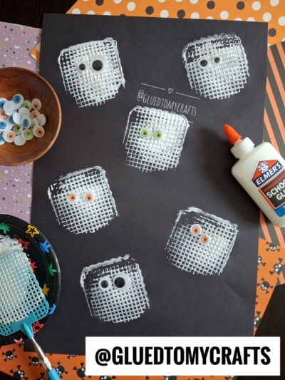 Six white ghost shapes with googly eyes are stamped on black paper using a fly swatter and white paint—perfect for Halloween Crafts for Kids. Glue, googly eyes, and Halloween-themed papers are nearby. The handle @GLUEDTOMYCRAFTS is displayed.