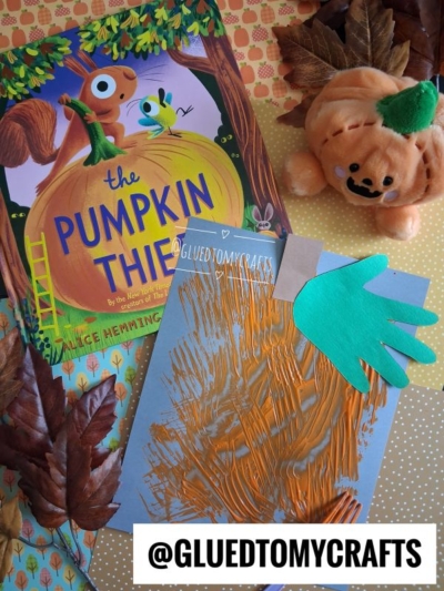 CHILDRENS pumpkin craft with a painted paper plate, green paper hand cutout, The Pumpkin Thief book, a pumpkin plush toy, fake autumn leaves, and the text @GLUEDTOMYCRAFTS at the bottom—a perfect addition to your collection of FALL CRAFTS FOR KIDS.