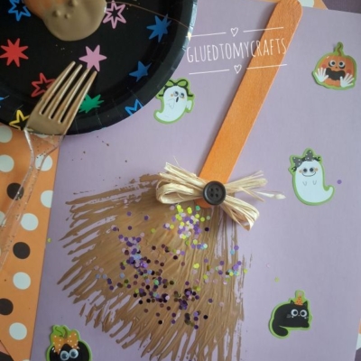 Childrens witch broom craft with a craft stick, ribbon, and glitter on purple paper, decorated with Halloween stickers. A chocolate-covered cookie and fork are on a starry black plate beside it. @GLUEDTOMYCRAFTS is printed below.