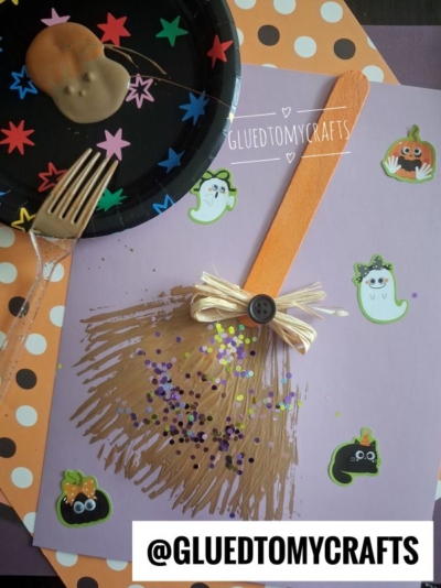 Childrens witch broom craft with a craft stick, ribbon, and glitter on purple paper, decorated with Halloween stickers. A chocolate-covered cookie and fork are on a starry black plate beside it. @GLUEDTOMYCRAFTS is printed below.