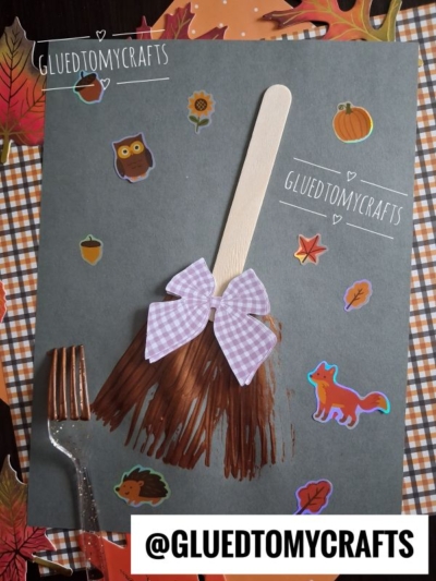 A fall-themed craft featuring a popsicle stick and brown paint shaped like a fork painted witch broom with a purple bow on dark paper, surrounded by autumn stickers and a fork, with @GLUEDTOMYCRAFTS text at the bottom.
