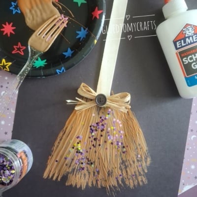 A popsicle stick and raffia broom craft decorated with a bow, button, and glitter sits on black paper. Nearby are glue, a fork with peanut butter, and a plate with colorful star stickers.