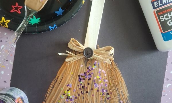 A popsicle stick and raffia broom craft decorated with a bow, button, and glitter sits on black paper. Nearby are glue, a fork with peanut butter, and a plate with colorful star stickers.
