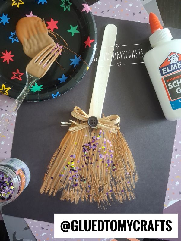 A popsicle stick and raffia broom craft decorated with a bow, button, and glitter sits on black paper. Nearby are glue, a fork with peanut butter, and a plate with colorful star stickers.