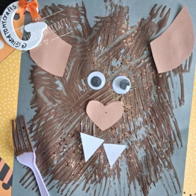 A bear face craft made with brown paint, googly eyes, and paper shapes on blue paper, with a purple fork and GLUED TO MY CRAFTS tags visible.