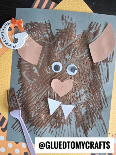 A bear face craft made with brown paint, googly eyes, and paper shapes on blue paper, with a purple fork and GLUED TO MY CRAFTS tags visible.