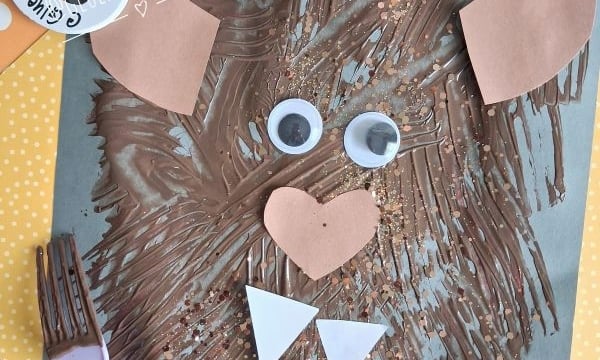 A bear face craft made with brown paint, googly eyes, and paper shapes on blue paper, with a purple fork and GLUED TO MY CRAFTS tags visible.