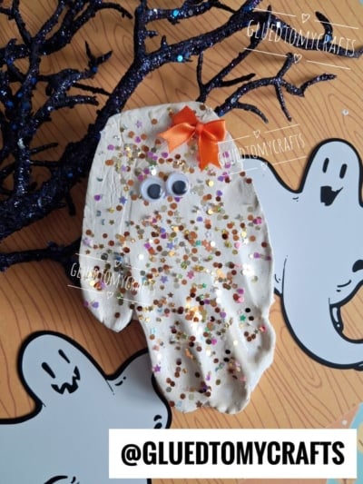 A handmade ghost craft with googly eyes, gold and orange glitter, and an orange bow sits on spooky paper with cartoon ghosts and a sparkly black branch—a perfect addition to your collection of Halloween Crafts for Kids. Text reads @GLUEDTOMYCRAFTS.