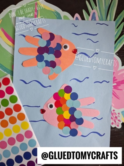 Two colorful fish crafts made from handprint cutouts and round stickers are shown on blue paper with wavy lines, displayed on a table with a floral background. These playful creations are perfect coding label crafts for organizing or identifying student projects. @GLUEDTOMYCRAFTS is written on the image.