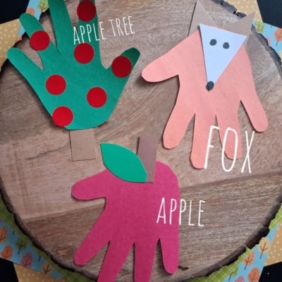 Three colorful hand-shaped paper crafts: a green apple tree with red dots, a red apple, and an orange fox face, all on a wooden surface with a @gluedtomycrafts label at the bottom.