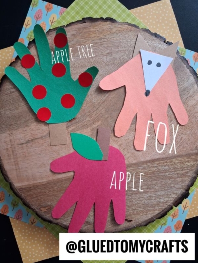 Three colorful hand-shaped paper crafts: a green apple tree with red dots, a red apple, and an orange fox face, all on a wooden surface with a @gluedtomycrafts label at the bottom.