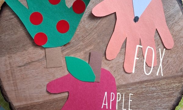 Three colorful hand-shaped paper crafts: a green apple tree with red dots, a red apple, and an orange fox face, all on a wooden surface with a @gluedtomycrafts label at the bottom.