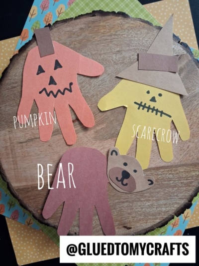 Three handprint crafts on a wooden background: an orange pumpkin with a jack-o-lantern face, a yellow scarecrow with a hat, and a brown bear with drawn-on features. Text labels each craft. @GLUEDTOMYCRAFTS at the bottom.