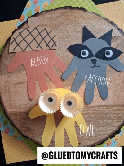 Three handprint crafts: an acorn, a raccoon, and an owl made with colored paper and cupcake liners, displayed on a wooden surface with patterned paper beneath. Labels name each craft.