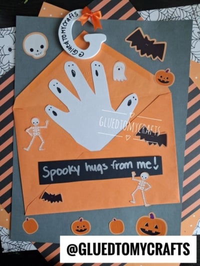 An orange envelope with a white handprint, labeled Spooky hugs from me! Decorated with Halloween stickers: skeletons, ghosts, pumpkins, bats, and a spider. This fun project is perfect for Halloween Crafts for Kids. @GLUETOMYCRAFTS is written at the bottom.
