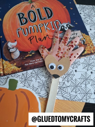 A handprint pumpkin craft with googly eyes lies on a Halloween-themed book called A Bold Pumpkin Plan—perfect for halloween story time—next to spiderweb-patterned paper and pumpkin cutouts.