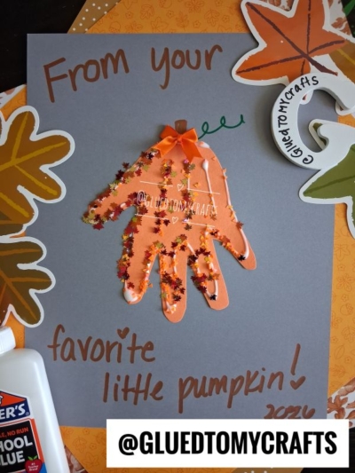 Handprint pumpkin craft on gray paper with “From your favorite little pumpkin!” written in brown. Surrounded by fall leaves, glue, and a pumpkin decoration, this sweet project is perfect for any FALL CRAFTS FOR KIDS collection. Features an orange handprint decorated with glitter and a bow.