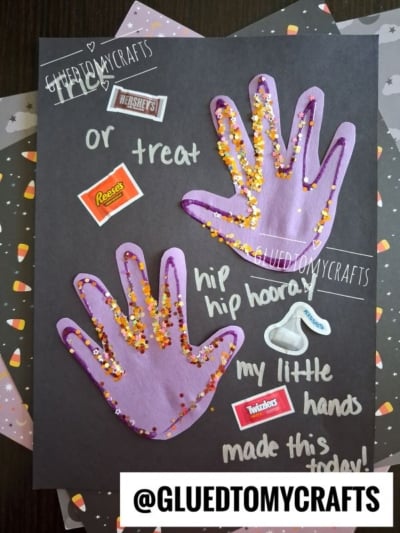A Halloween craft shows two purple handprints with glitter on black paper, surrounded by candy stickers and phrases like hip hip hooray! and my little hands made this today! Perfect for your next session of Halloween Crafts for Kids, this adorable keepsake also features the username @GLUEDTOMYCRAFTS at the bottom.