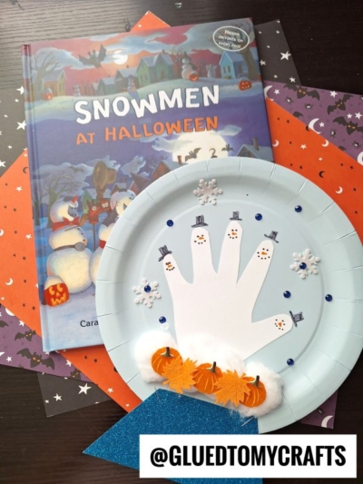 A children’s book titled Snowmen at Halloween sits next to a charming paper plate craft featuring a handprint snowman inside a sparkly snow globe, along with mini pumpkin cutouts and snowflake stickers. The Instagram handle @GLUEDTOMYCRAFTS is at the bottom.
