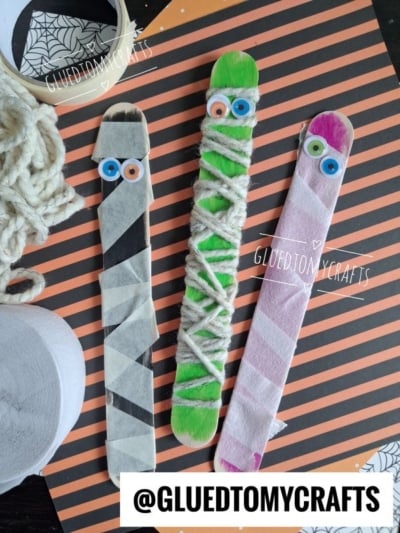 Three craft sticks decorated as mummies with googly eyes, yarn, and tape, placed on a black-and-orange striped background with rolls of tape and yarn nearby—a fun and easy idea for Halloween Crafts for Kids.