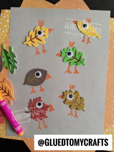 A gray sheet features FOAM LEAF STICKER CRAFTS with googly eyes and drawn wings and legs, making them look like birds. Colorful markers and leaf-patterned paper are in the background. @GLUEDTOMYCRAFTS at the bottom.