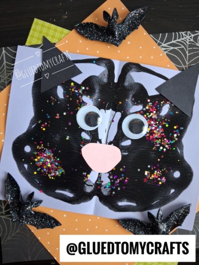 A symmetrical black paint blot craft resembling a cat face on light purple paper, featuring googly eyes, a pink nose, and paper ears. Perfect for halloween story time, it is surrounded by glittery black bat cutouts on an orange background with a spiderweb pattern.