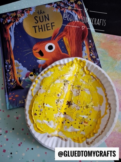 A children’s book titled The Sun Thief is next to a paper plate art project covered in yellow and white paint, on a pastel dotted surface—perfect inspiration for summer craft ideas. The image is watermarked with @GLUEDTOMYCRAFTS.