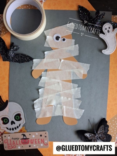 A mummy craft made from brown paper and white tape is surrounded by Halloween decorations: black bats, a ghost, a skull, a butterfly, tape, and an orange Happy Halloween card. Perfect for anyone searching for fun Halloween Crafts for Kids, @GLUETOMYCRAFTS is visible.