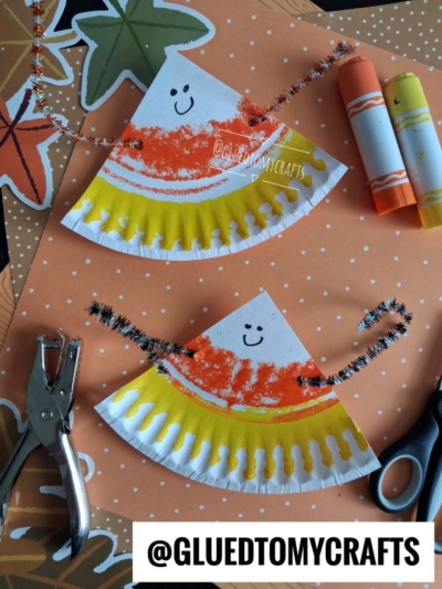 Two paper plate candy corn crafts with smiling faces, decorated with crayon and pipe cleaners, are placed on a dotted orange surface with markers, scissors, and a hole punch nearby.