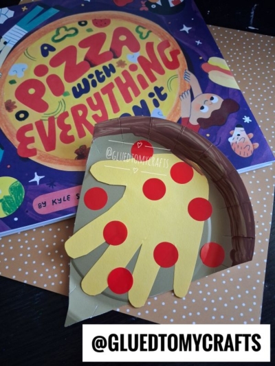 A children’s book titled A Pizza With Everything On It is next to a handmade pizza slice craft made from colored paper shaped like a hand. Red dot pepperoni and a brown paper crust are on the craft, making it one of our favorite summer craft ideas. @GLUEDTOMYCRAFTS.
