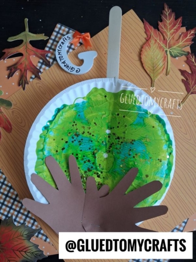 A fall-themed craft shows a paper plate with green and blue paint splat, brown paper hand cutouts, a popsicle stick, faux leaves, and a small painter’s palette, all arranged on an autumn-patterned background—perfect inspiration for making your own caramel apple art this season.