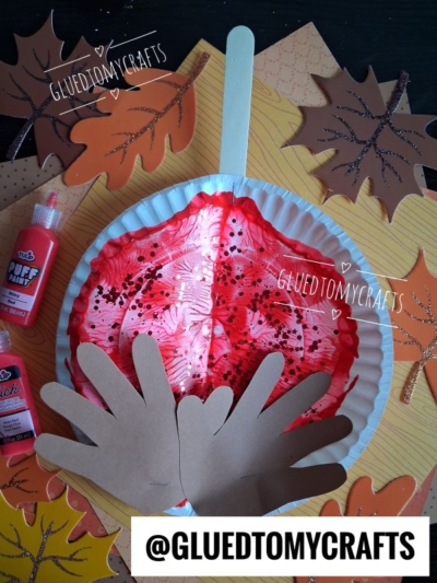 A red painted paper plate craft shaped like a Native American headdress, featuring brown paper hand cutouts as feathers, surrounded by paper leaves, craft supplies, and a playful paint splat caramel apple accent. @GLUEDTOMYCRAFTS is displayed at the bottom.