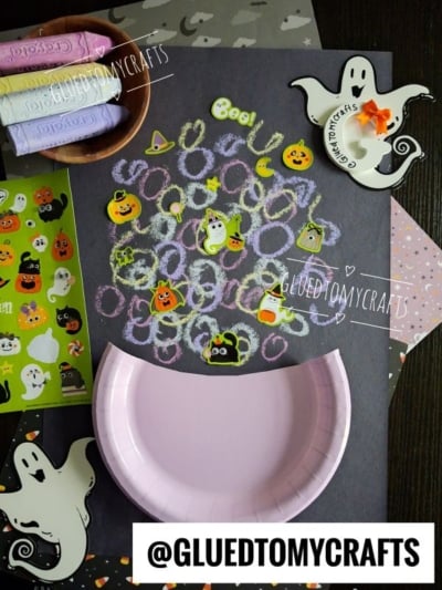 A Halloween-themed craft scene with a half paper plate, ghost and pumpkin stickers, pastel crayons, and various decorated papers on a table—perfect inspiration for Halloween Crafts for Kids. The words @GLUEDTOMYCRAFTS appear on the image.